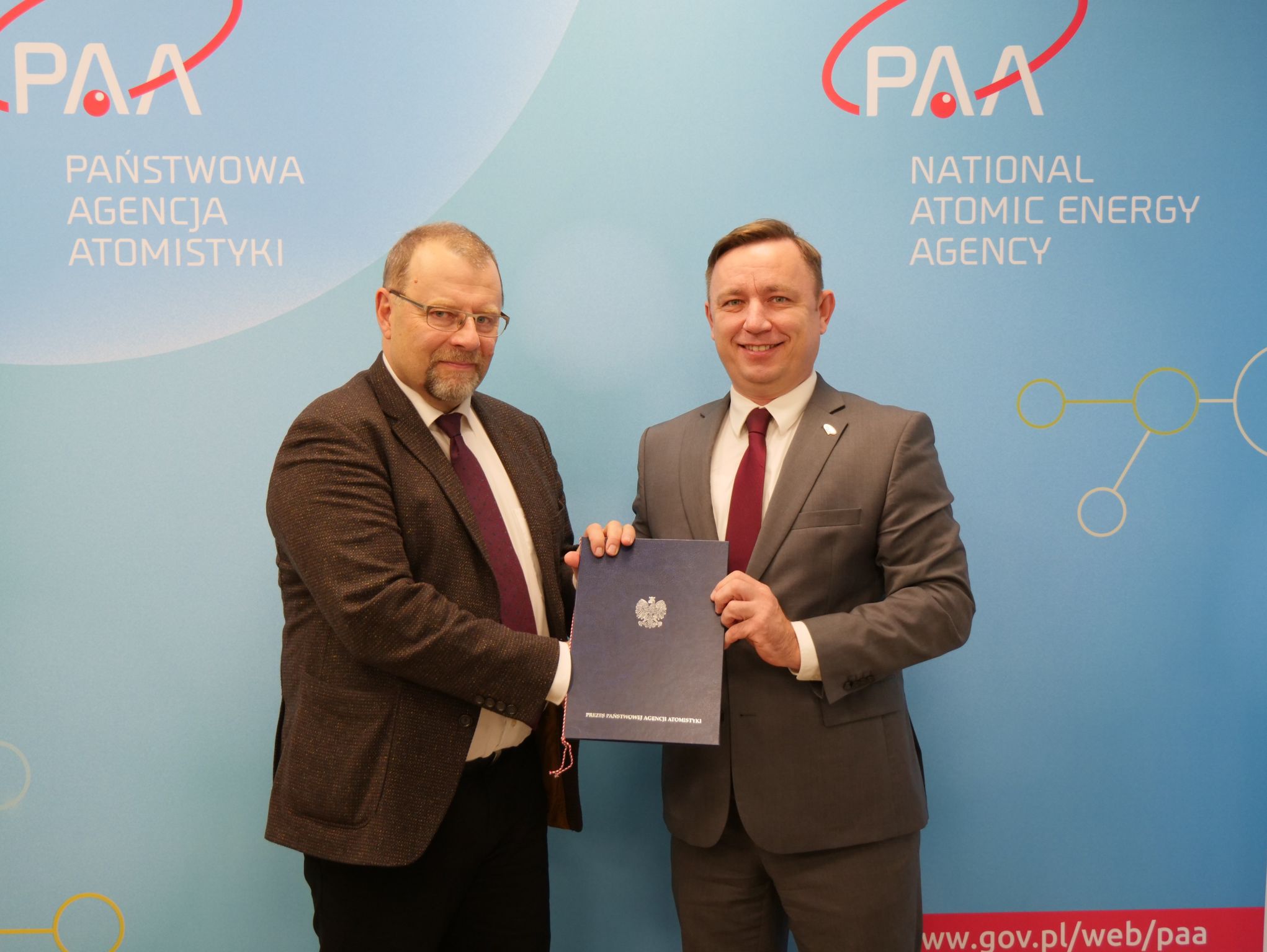 Paweł Smoleń (left hand side) together with somebody from the holding a document in front of the Państwowa Agencja Atomistyki (PAA) banner recognizing Kiwa Nuclear AB, represented in Poland by Kiwa Poland, as an expert organization qualified to carry out assessments for the application process of Poland’s first nuclear power plant 