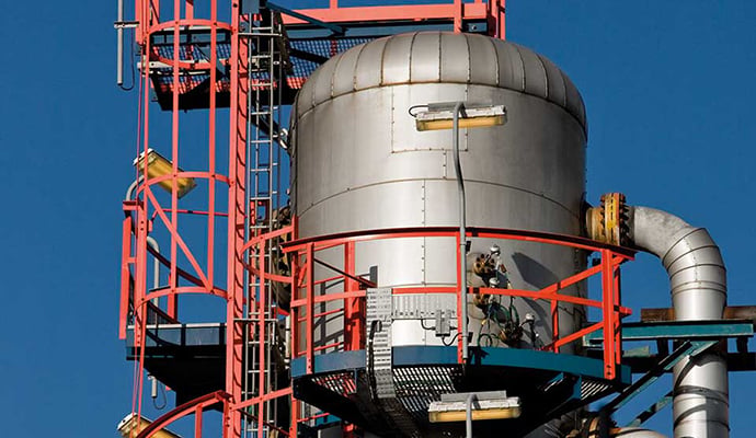 Industriell anläggning med en stålcistern omgiven av röda och blå metallramar och stegar under klarblå himmel.