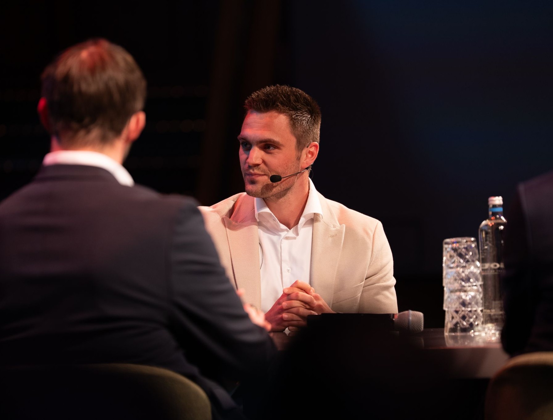 Bart Scholten, Business Unit Manager for Cyber Security, in a beige blazer speaking into a headset microphone at a conference table, engaged in conversation with another person, with water bottles visible on the table