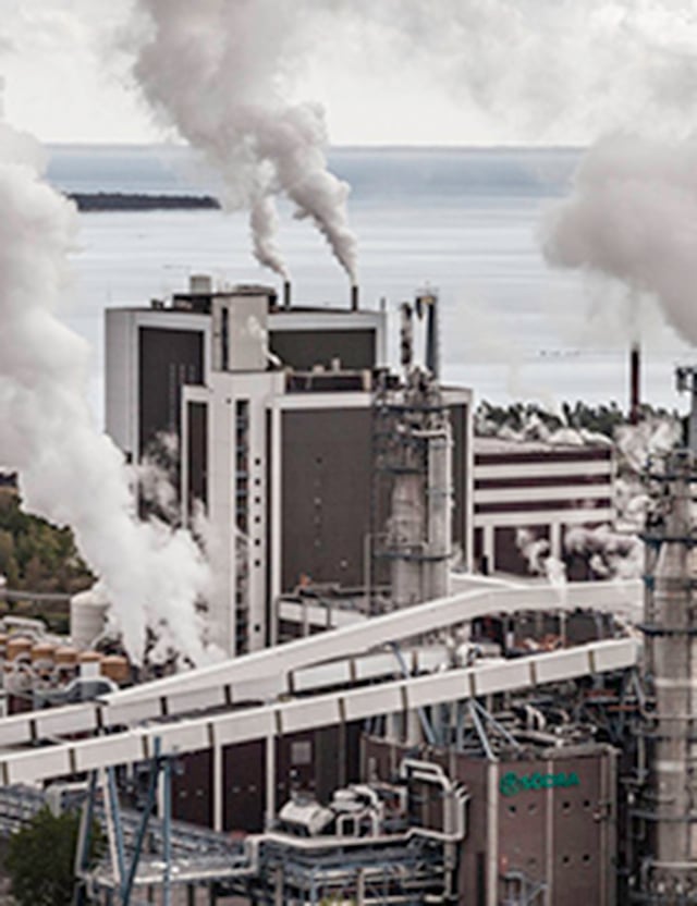 Industribyggnad med stora skorstenar som släpper ut vit rök nära en kustlinje.
