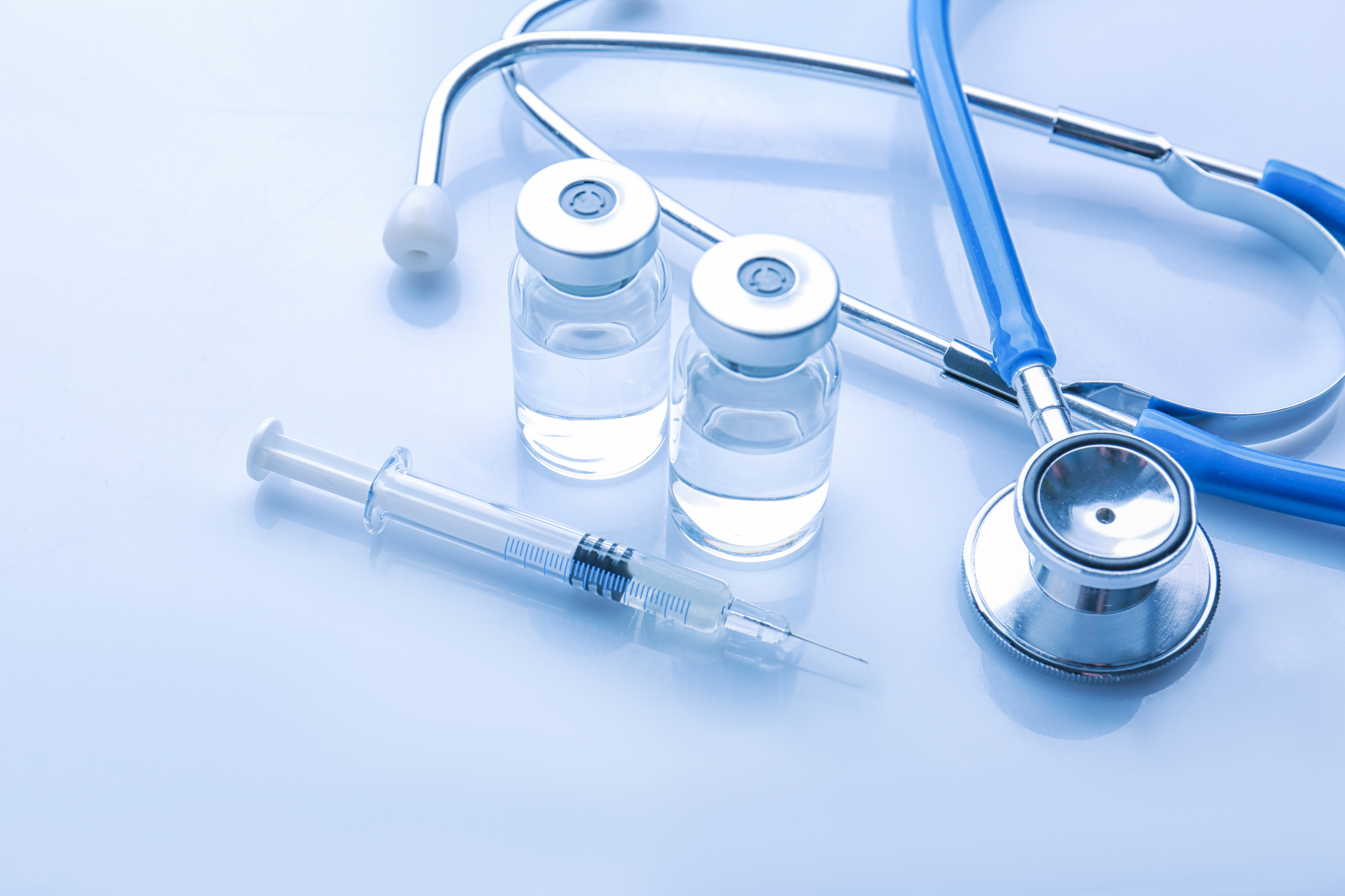 Medical supplies including a stethoscope, syringe, and two vaccine vials on a light blue surface.