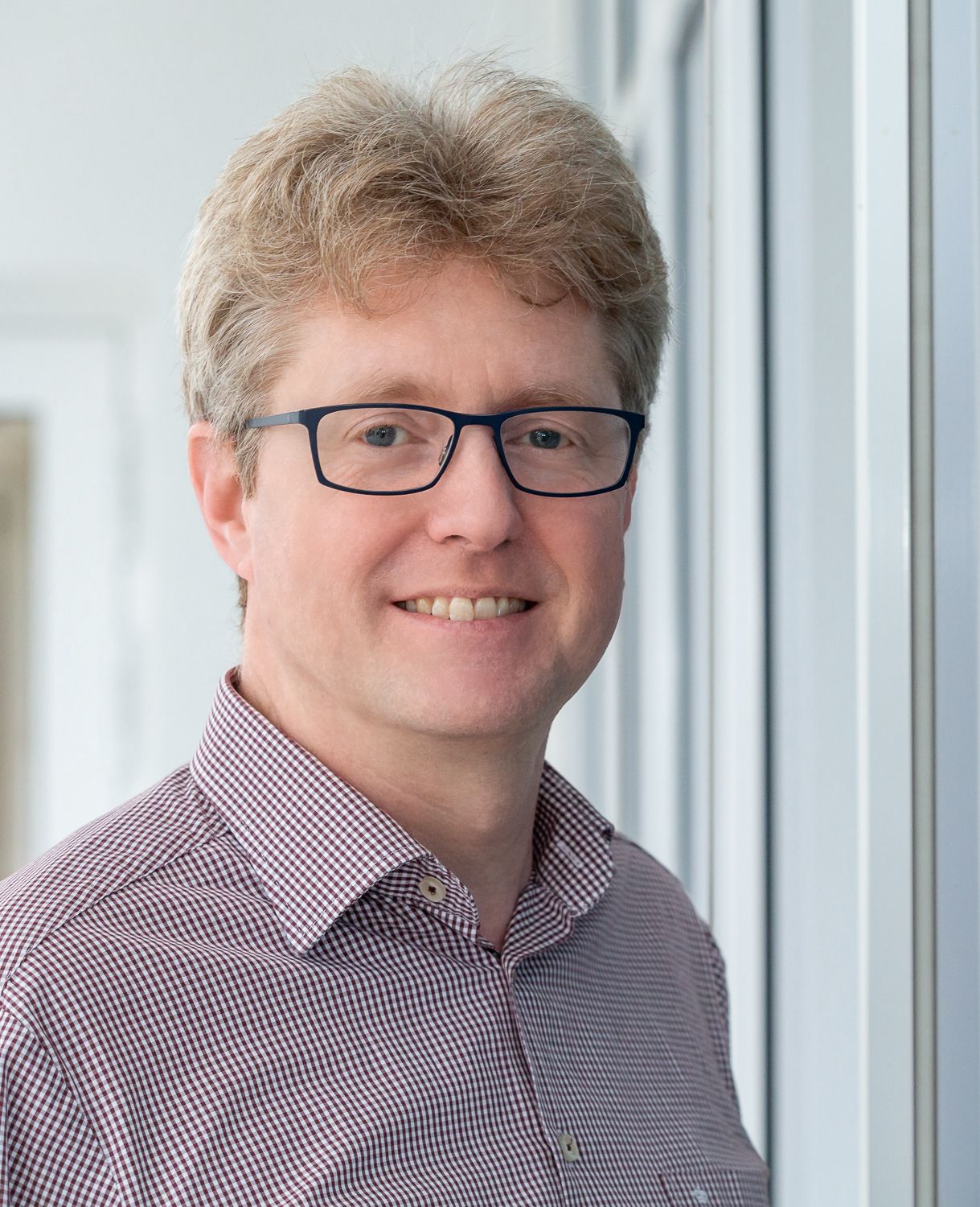 Carsten Rieck, International Business Development Manager for Construction and Infrastructure, smiling with glasses wearing a checkered shirt, standing indoors by a window