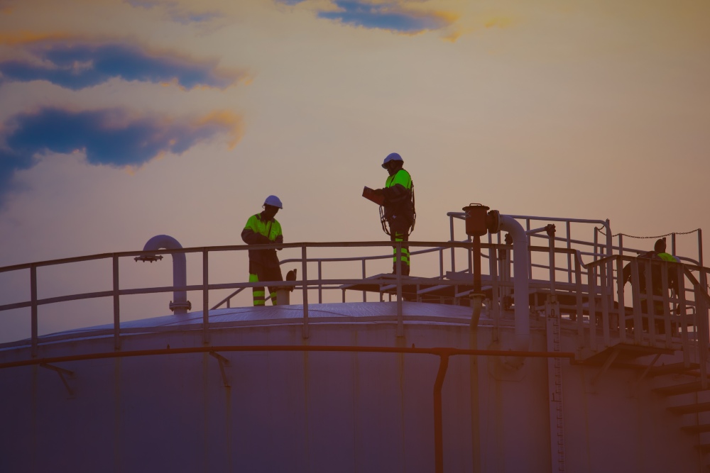 Workers in high-visibility safety gear standing on top of an industrial storage tank at sunset.
