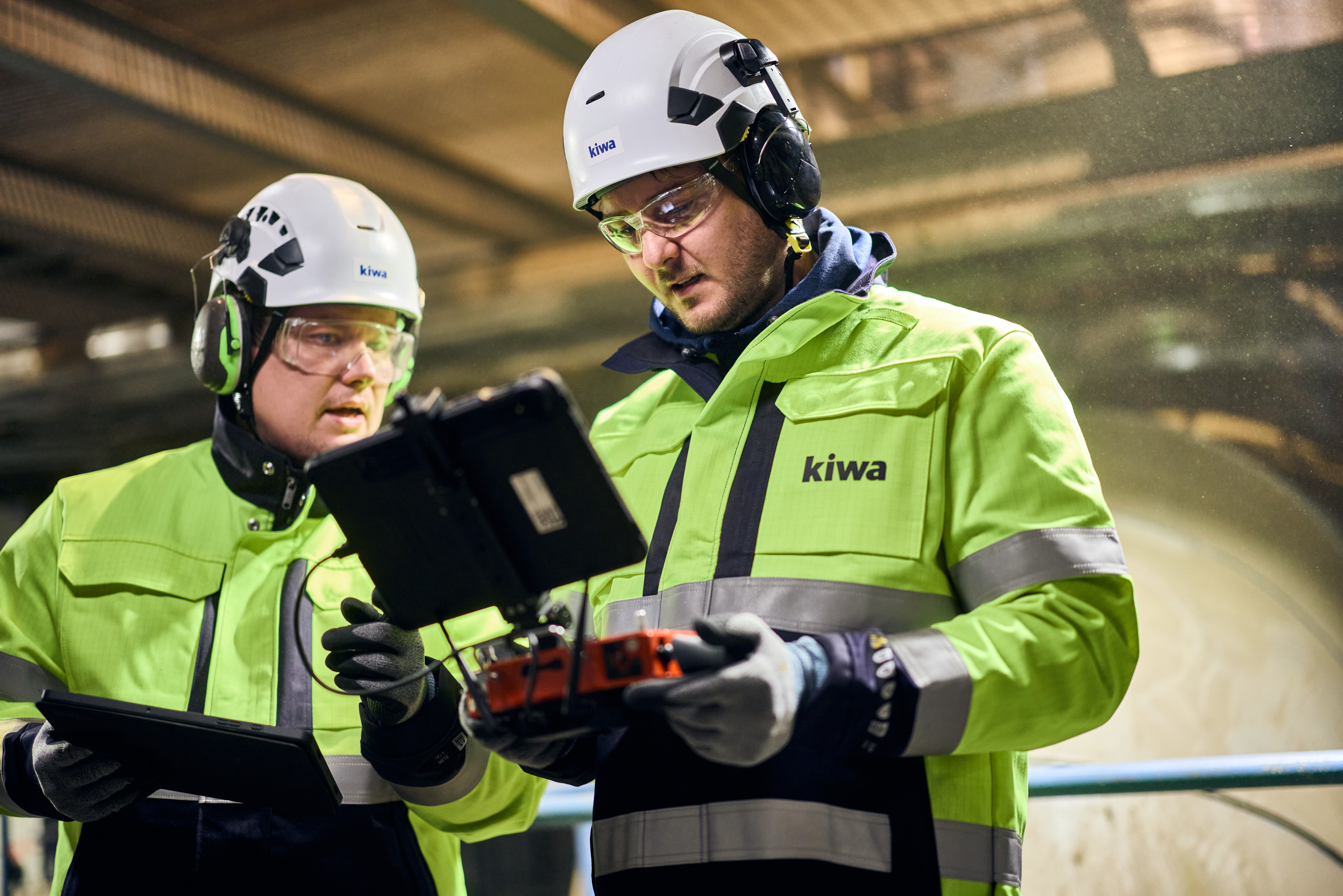 Two workers in high-visibility Kiwa safety jackets and helmets inspecting equipment with a tablet and control device in an industrial setting.