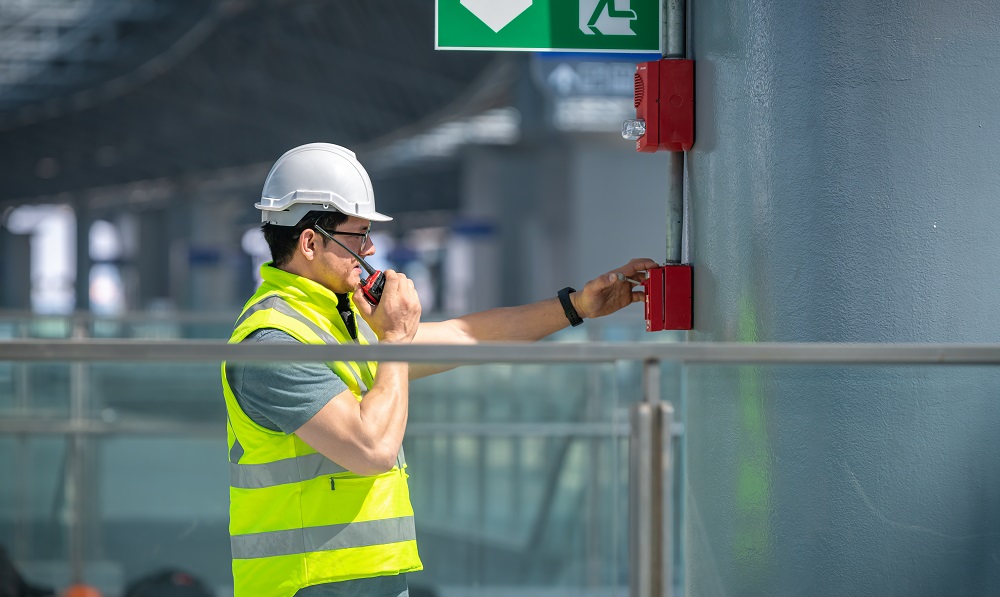 Kiwa auditor in a hard hat and high-visibility vest operating a wall-mounted fire alarm call point indoors.