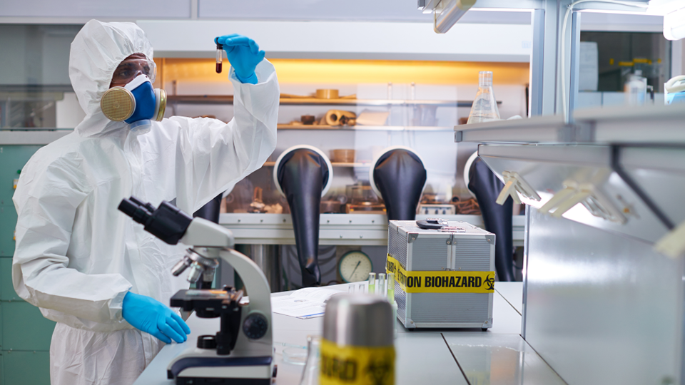 Person i skyddsdräkt och gasmask håller ett provrör i ett laboratorie med mikroskop, biohazard-märkning och annan forskningsutrustning. Hur det kan se ut när man arbetar med farliga ämnen.