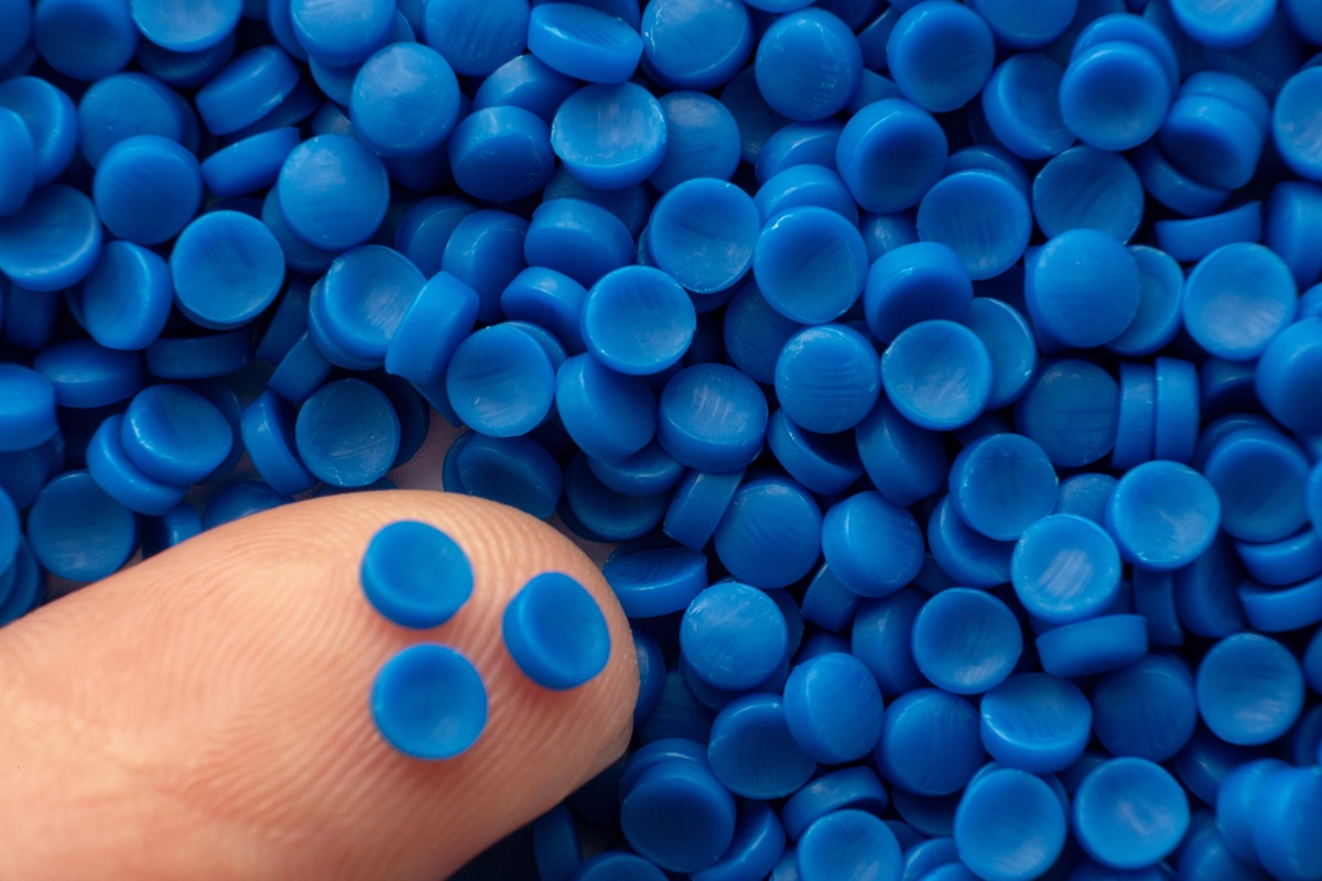 Close-up van een vinger met kleine blauwe kunststof korrels op de huid, omringd door een grote hoeveelheid blauwe pellets.