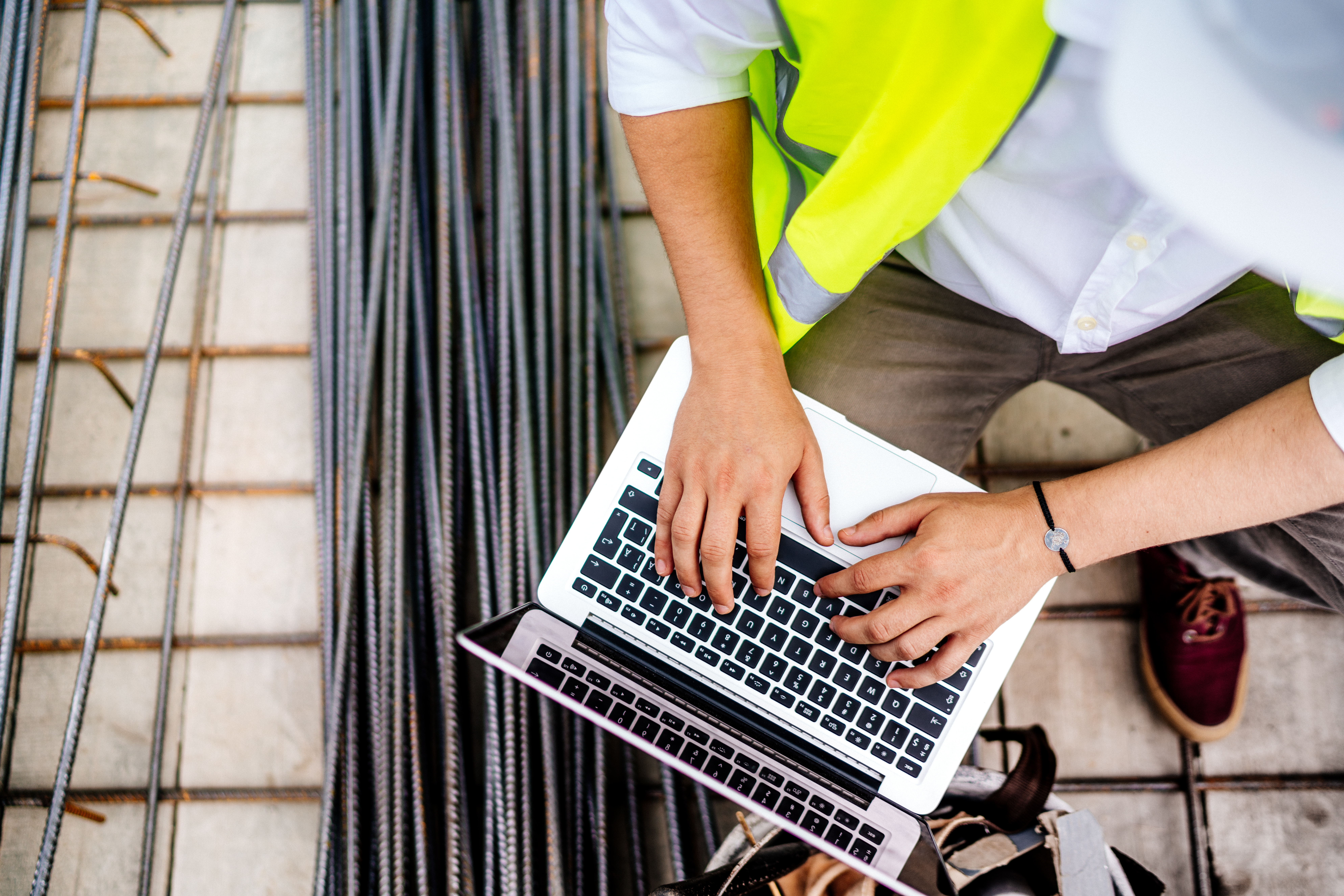 Kiwa employee in a safety vest using a laptop beside bundled steel rebar at a building site.