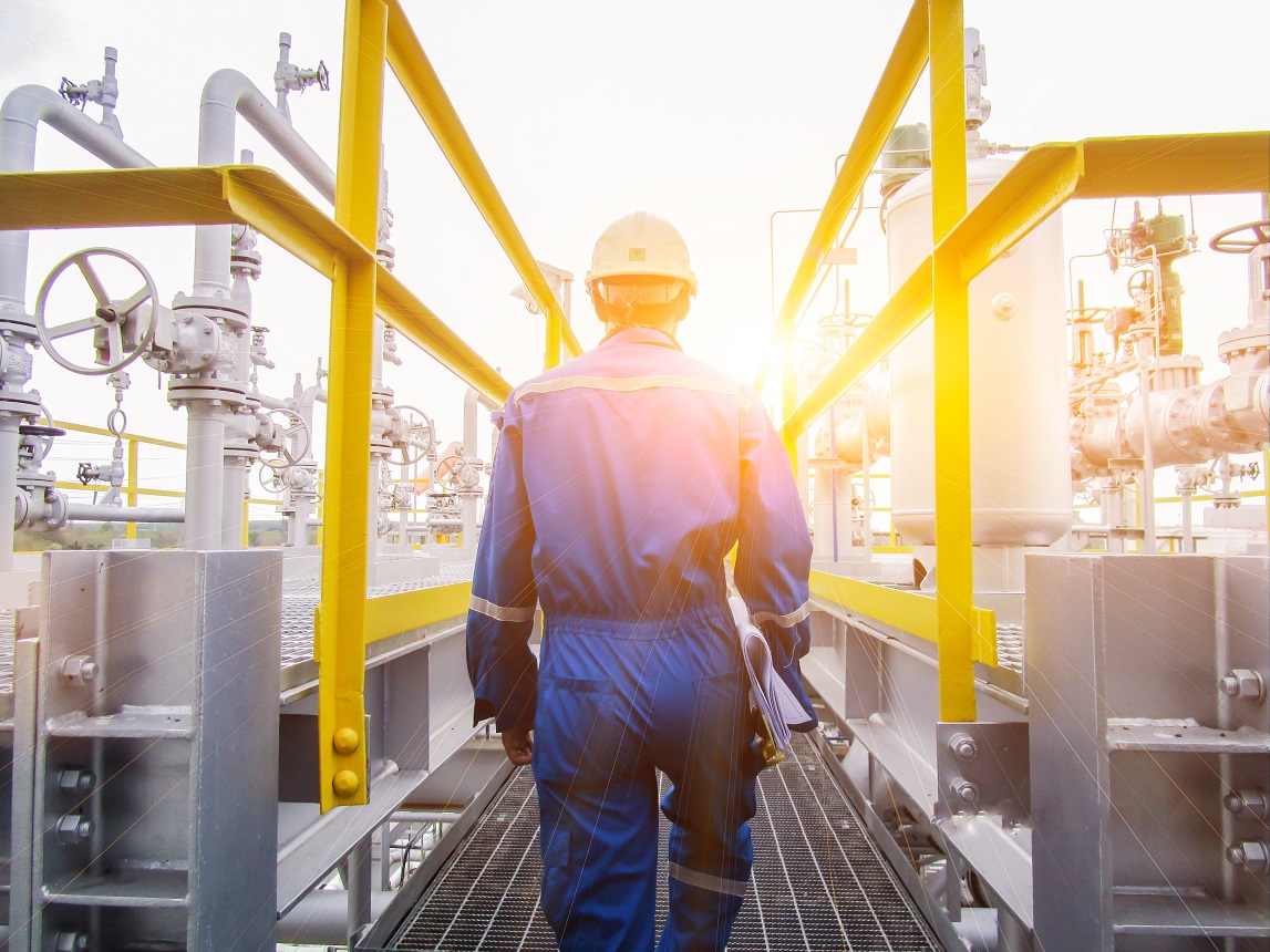 A technician in blue coveralls and a hard hat is walking through an industrial plant with pipes, valves and yellow railings
